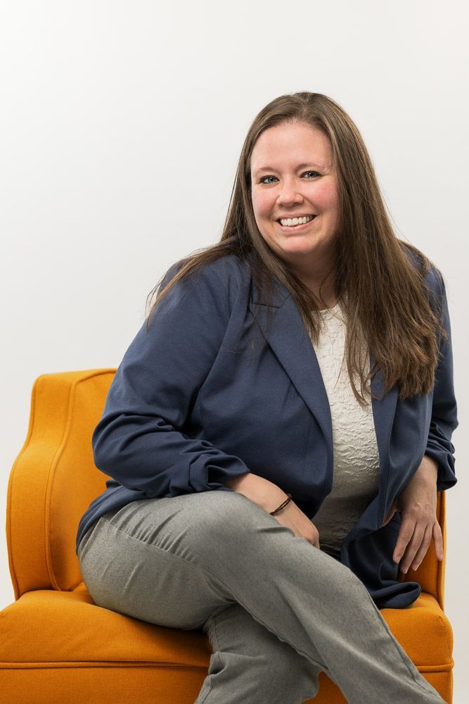 Woman with long brown hair, wearing a blue blazer and grey pants, smiles while sitting in an orange chair.
