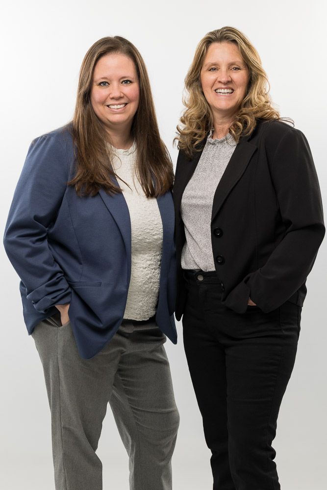 Two women in business attire, smiling, posing together in front of a white backdrop.