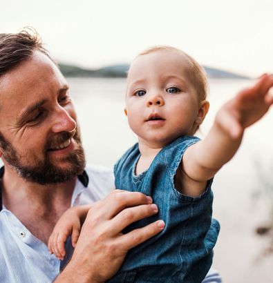A man smiles while holding a baby who reaches out, with a body of water in the background.