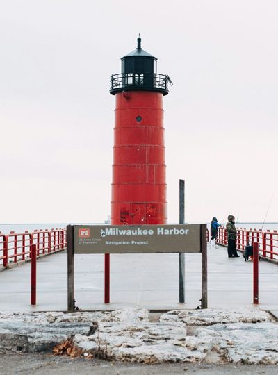 Red lighthouse at Milwaukee Harbor with a sign and people on a pier, under a cloudy sky.