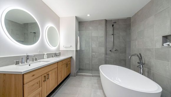 Modern bathroom with double vanity, round lit mirrors, gray tile shower, and freestanding tub.