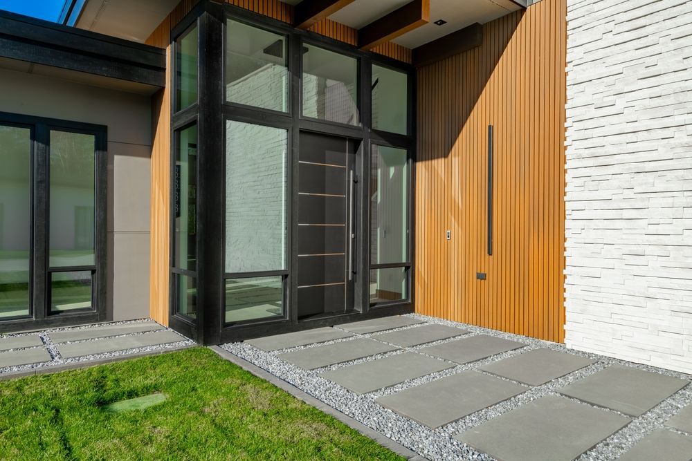Modern house entrance with black-framed glass doors, wood siding, white brick wall, and stone path by grass
