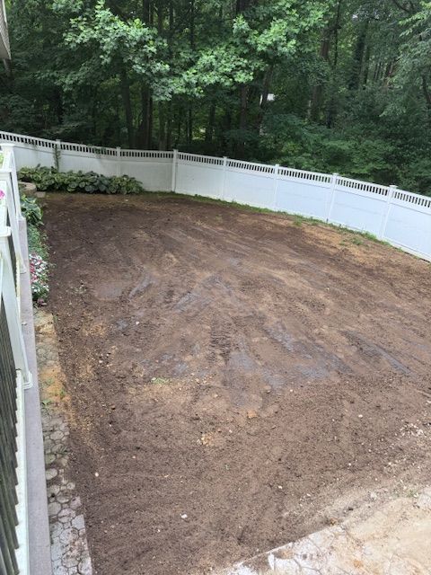 Freshly leveled dirt yard enclosed by a white fence beside a wooded area
