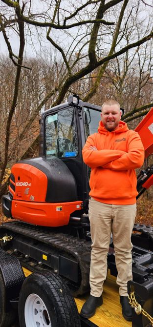 Man in orange hoodie stands beside an orange compact excavator in a wooded area.