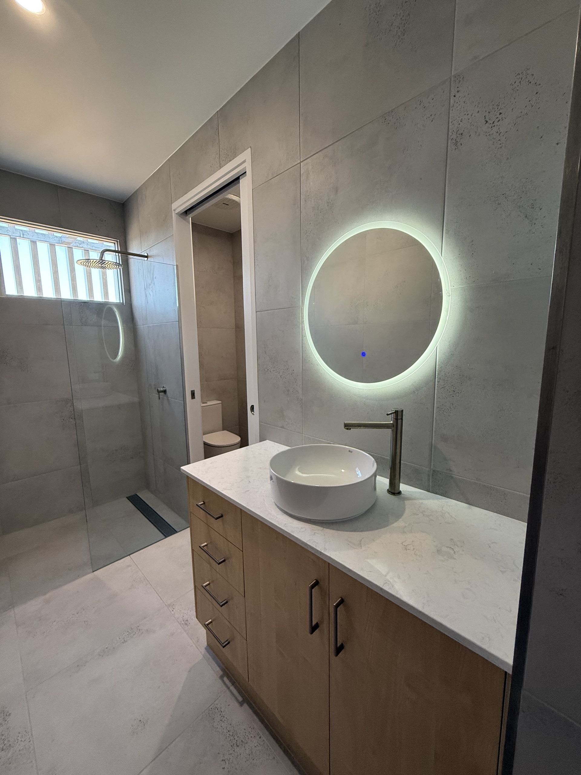 Modern bathroom with vanity, round mirror, sink, and walk-in shower with glass blocks — Coral Coast Building & Management in Cannon Valley, QLD 