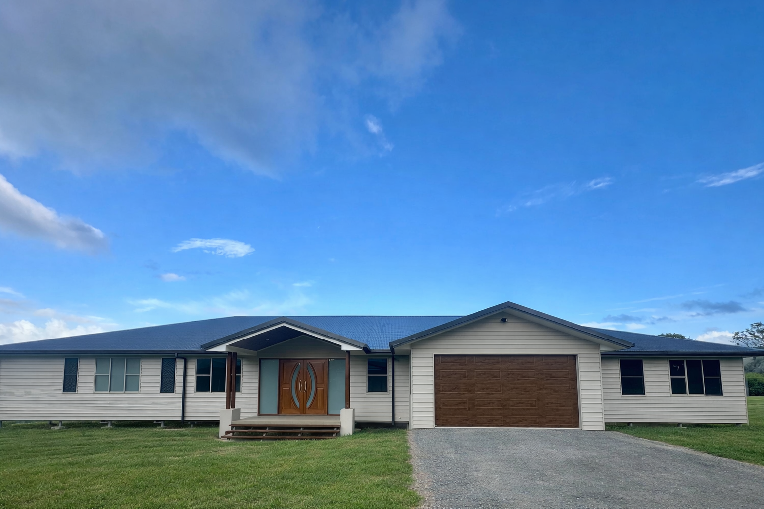 Single-story house with blue roof, white siding, brown garage door, and a gravel driveway under a blue sky — Coral Coast Building & Management in Cannon Valley, QLD