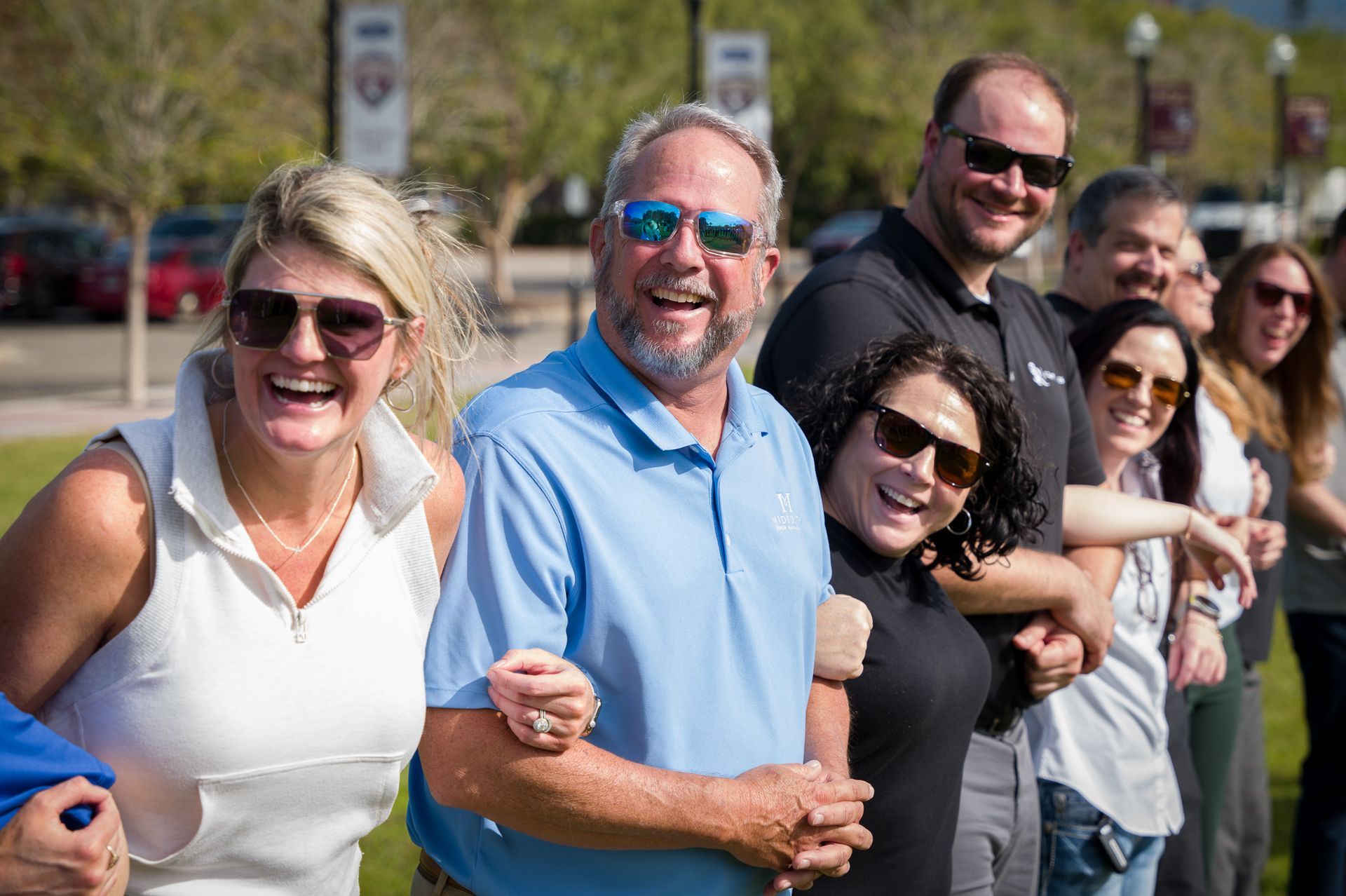 Smiling people standing arm in arm outdoors on a sunny day, with others lined up behind them