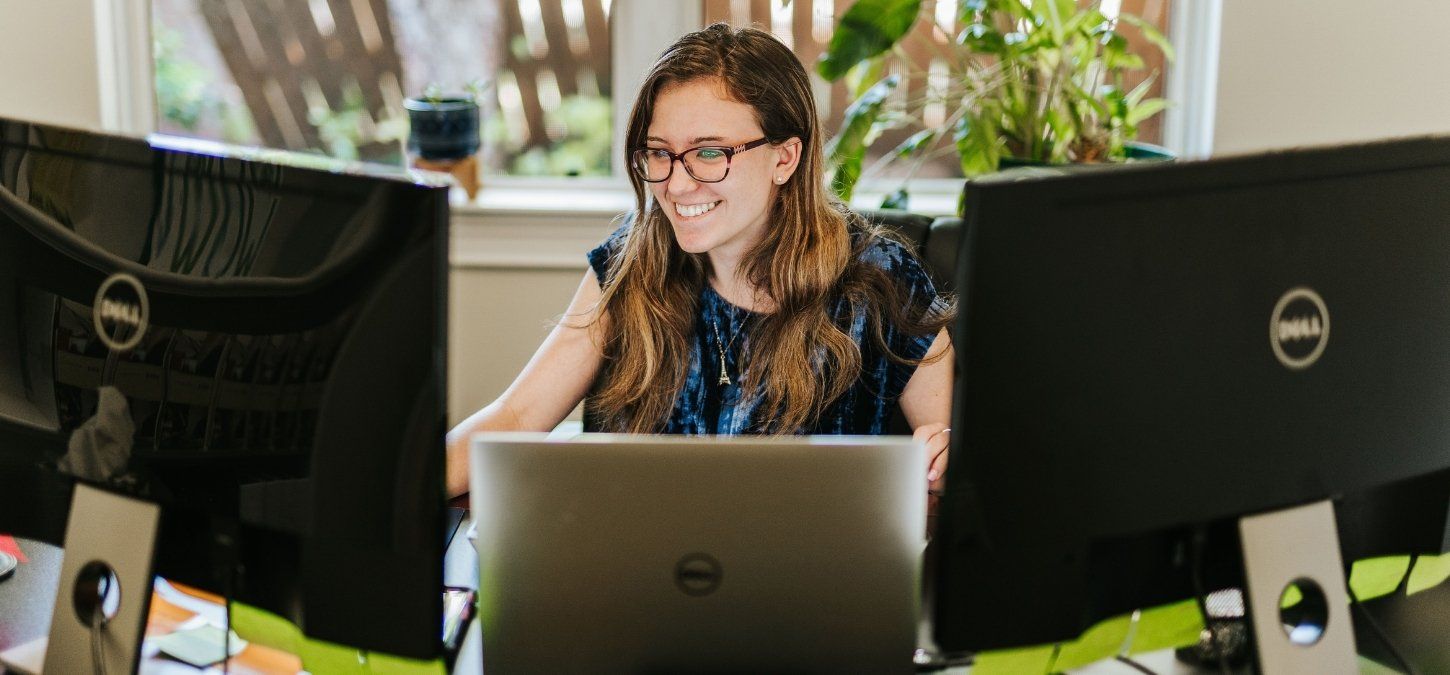 Woman smiling at a desk with two monitors and a laptop in a bright office