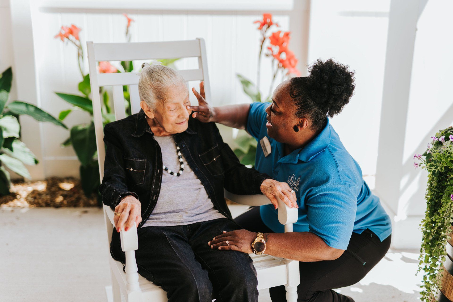 Caregiver helping an older adult sit in a chair on a bright porch garden area