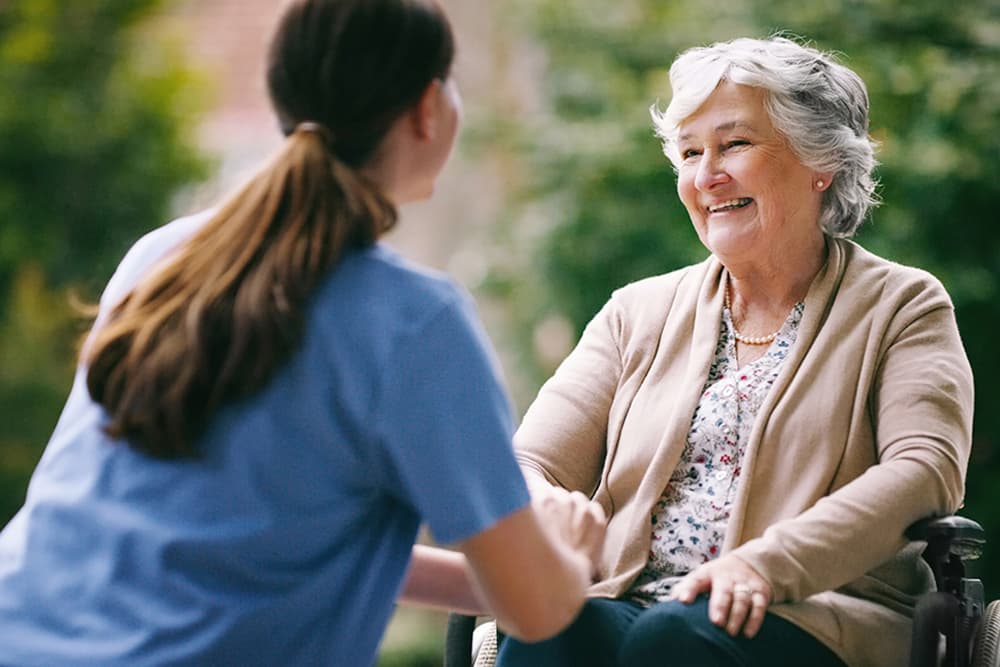 Woman in blue speaking with smiling elderly woman in a wheelchair outdoors.