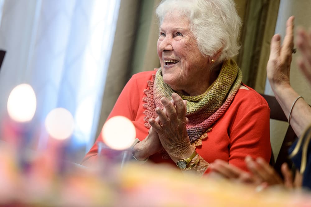 Smiling elderly woman in a red top clapping at a table with soft blurred lights in the foreground