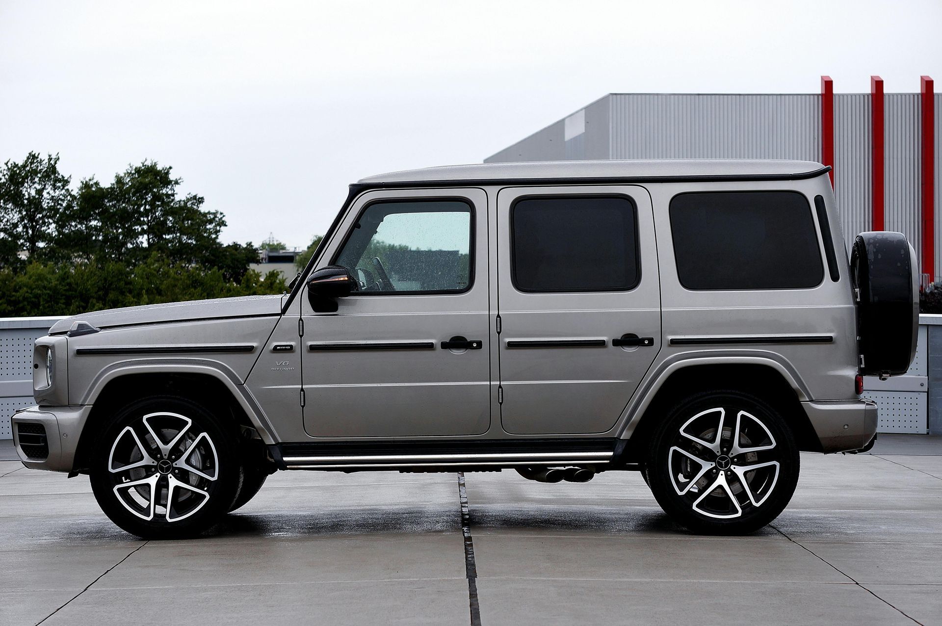 A side view of a silver Mercedes-Benz G-Wagon parked on a concrete surface against an industrial building backdrop.