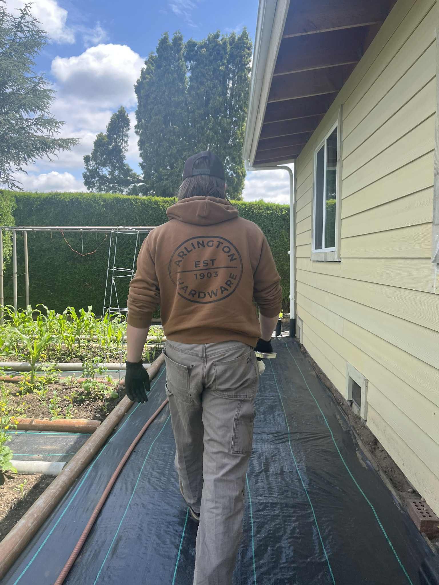 Person walking along a garden path; brown hoodie, light pants. Beside a yellow house, a garden on the left.