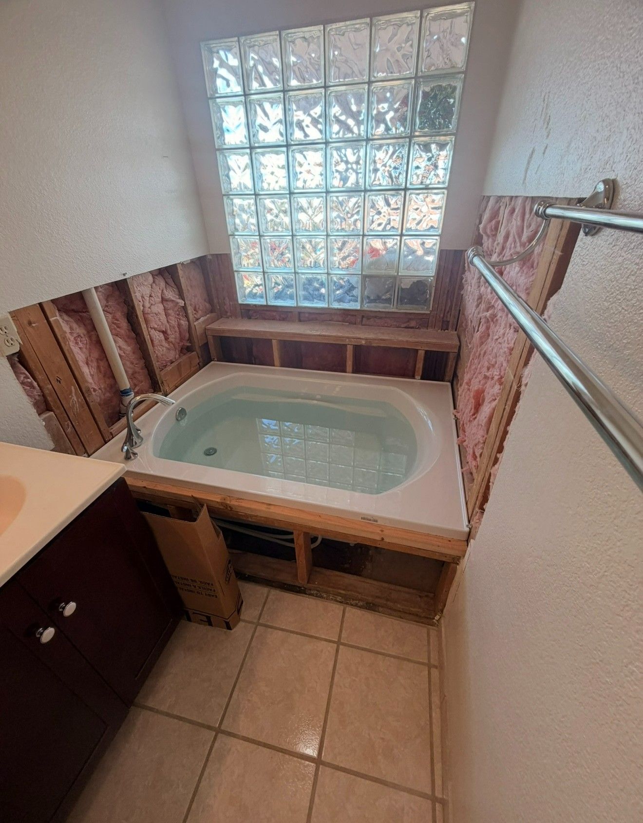 Bathroom with a built-in bathtub under a glass-block window, surrounded by exposed wall studs and tile floor.