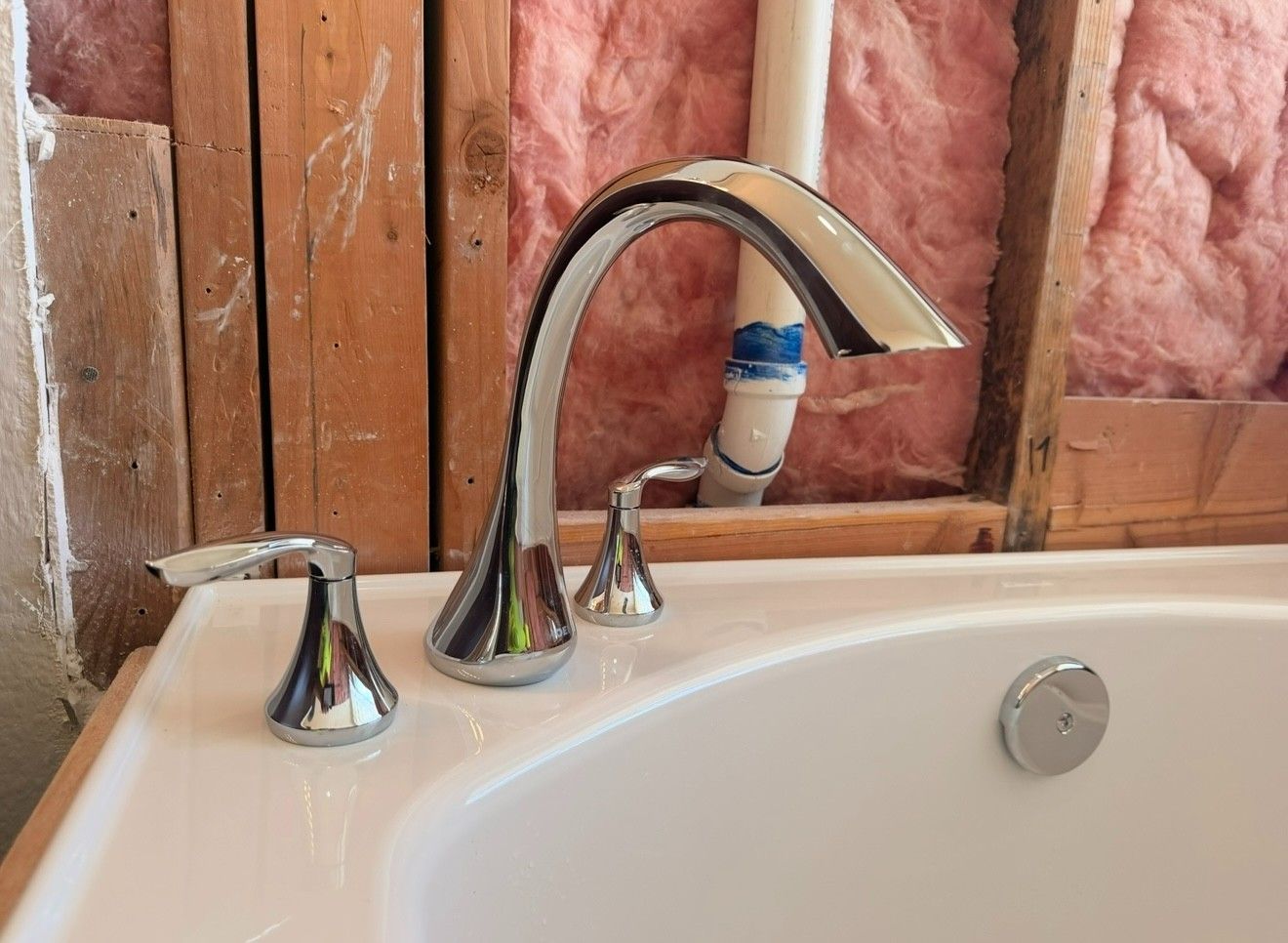 Chrome bathtub faucet and handles in front of exposed wall studs with pink insulation