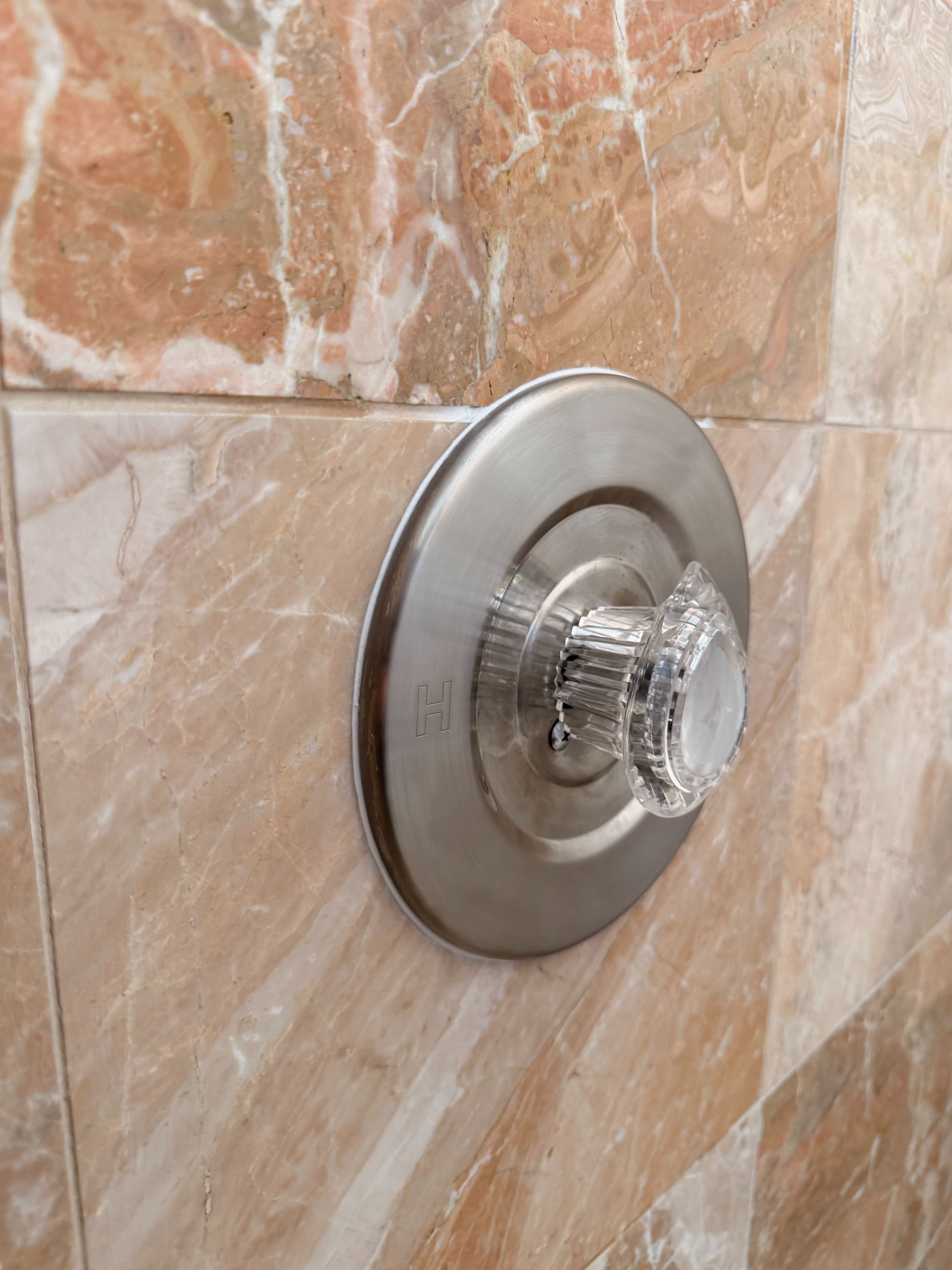 Chrome shower control knob on a beige tiled wall, with a clear plastic cover over the handle