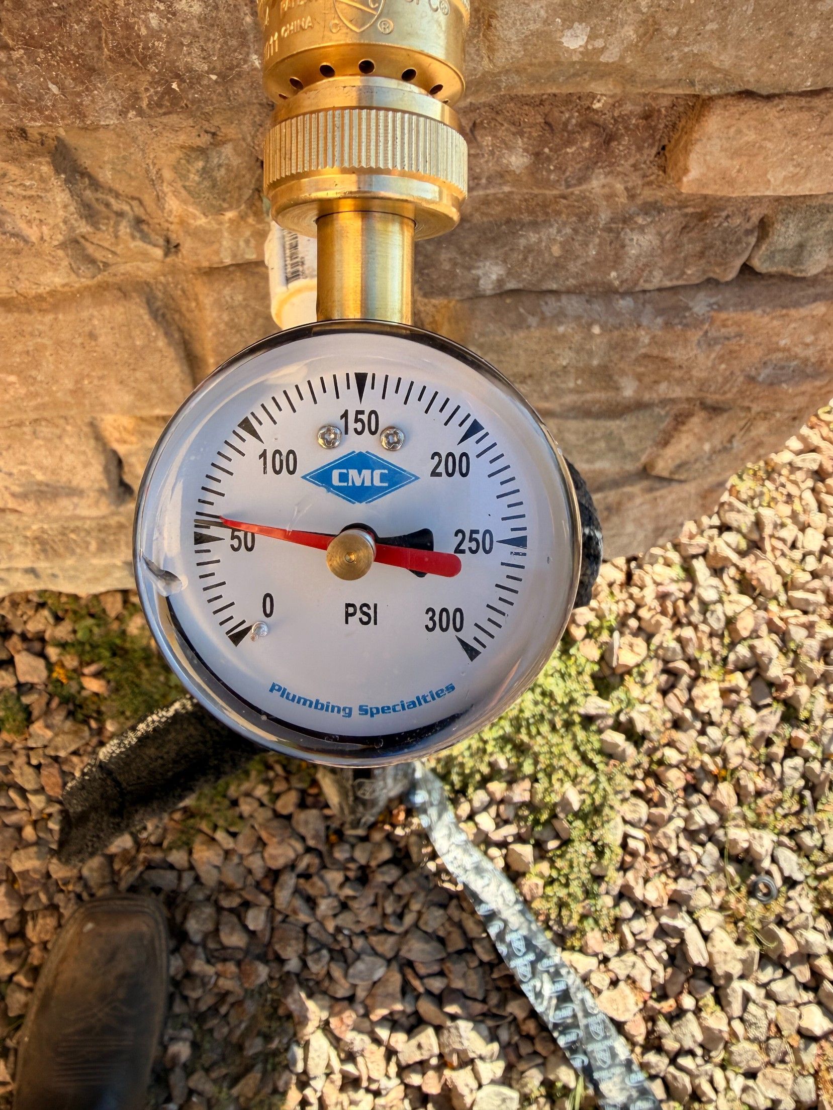 Close-up of a PSI pressure gauge reading around 250, attached to a pipe against a rock wall.