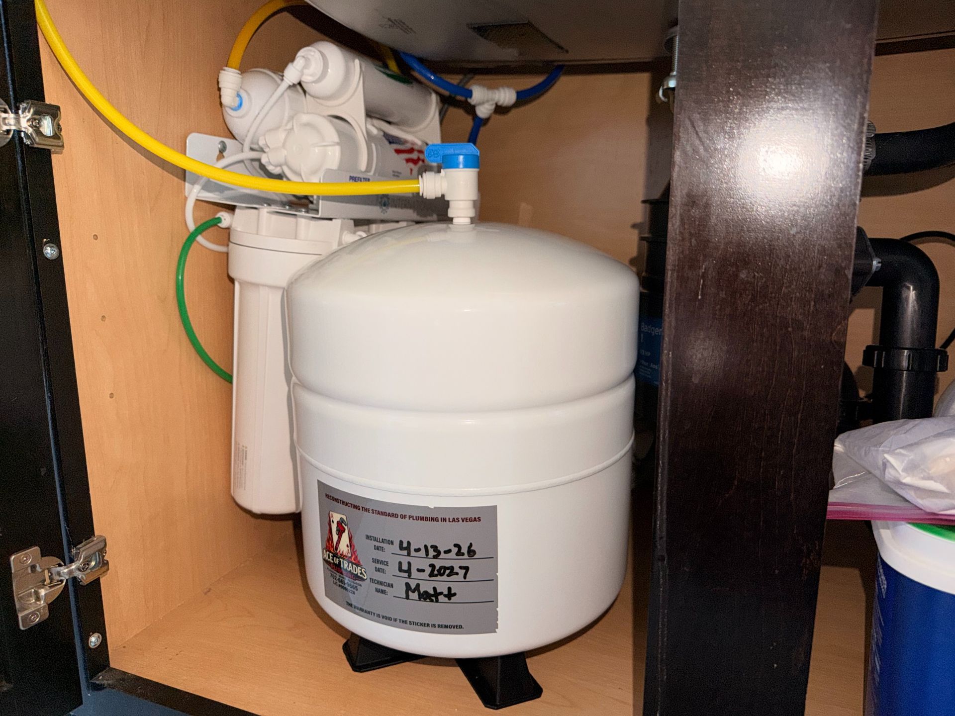 Close-up of a white pressure tank and plumbing in a cabinet, with colored hoses and pipes.