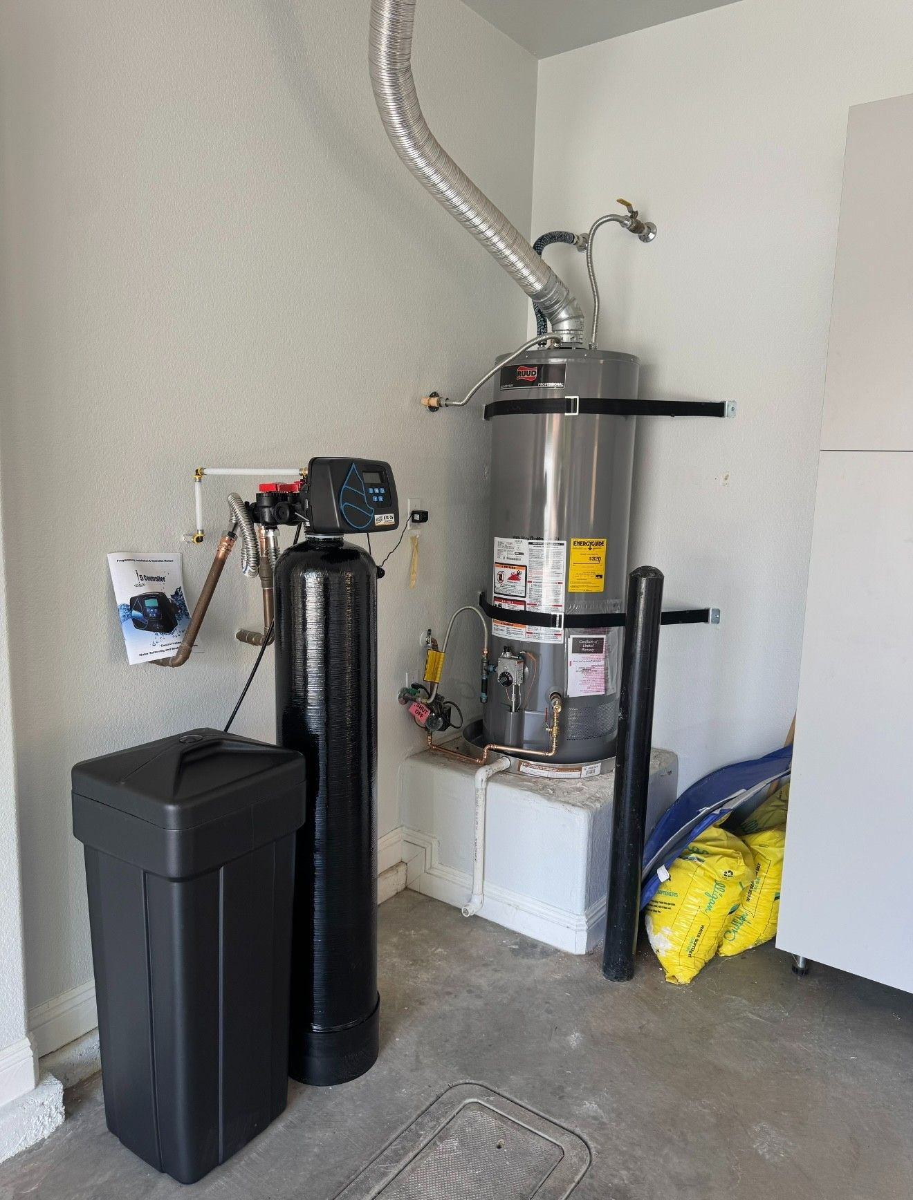 Garage corner with water heater, black tank, trash bin, and cleaning supplies on concrete floor