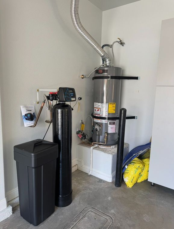 Garage corner with water heater, black tank, trash bin, and cleaning supplies on concrete floor