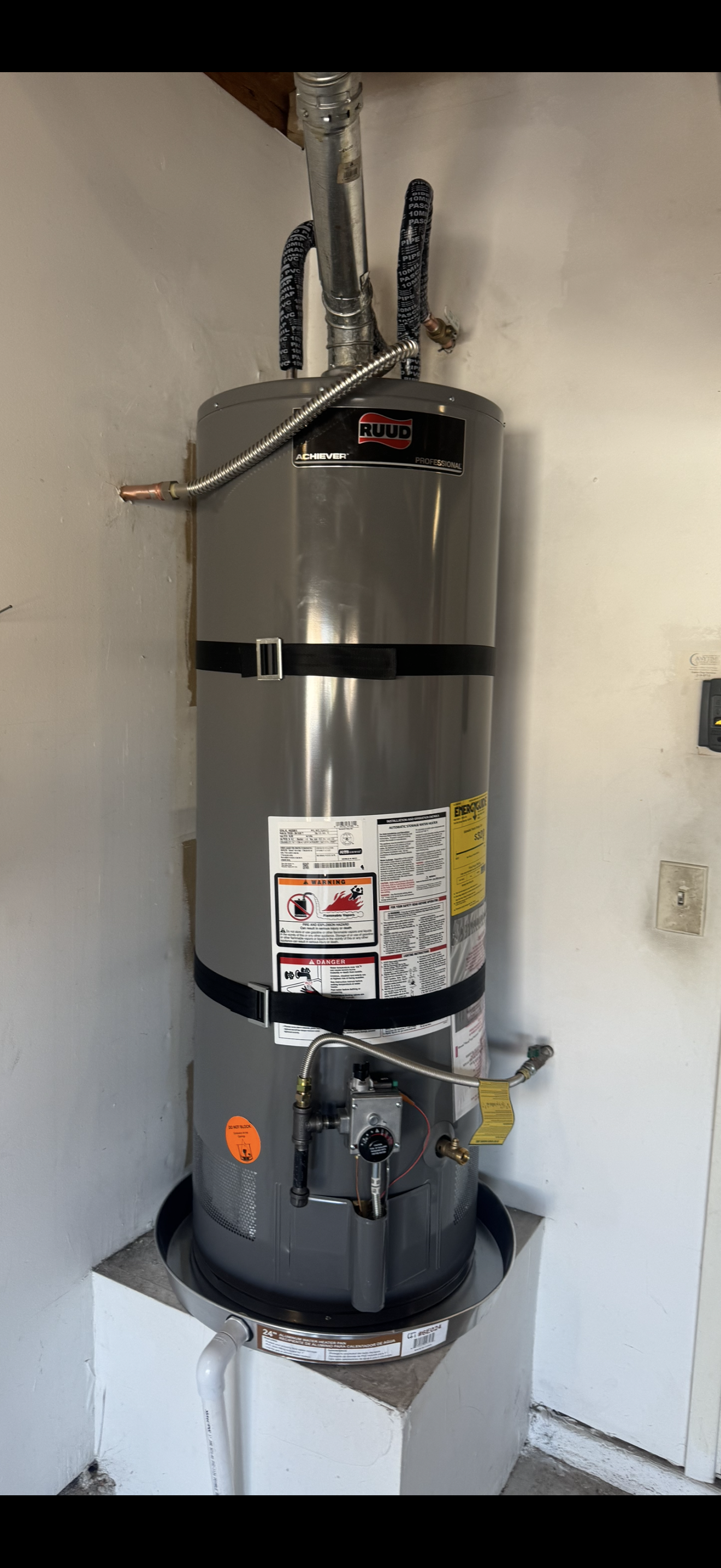 Tall silver water heater with pipes and labels in a utility closet, mounted above a black base pan
