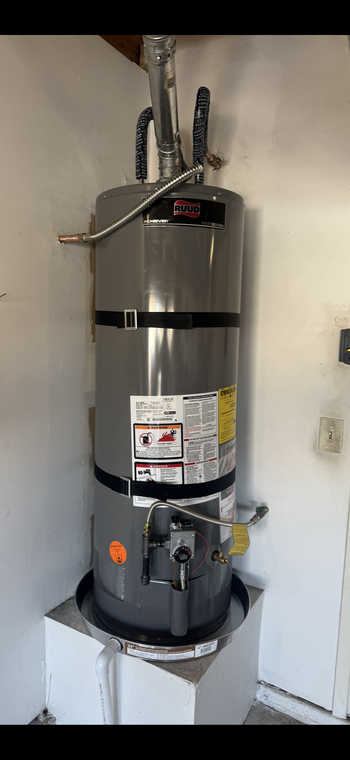 Tall silver water heater with pipes and labels in a utility closet, mounted above a black base pan