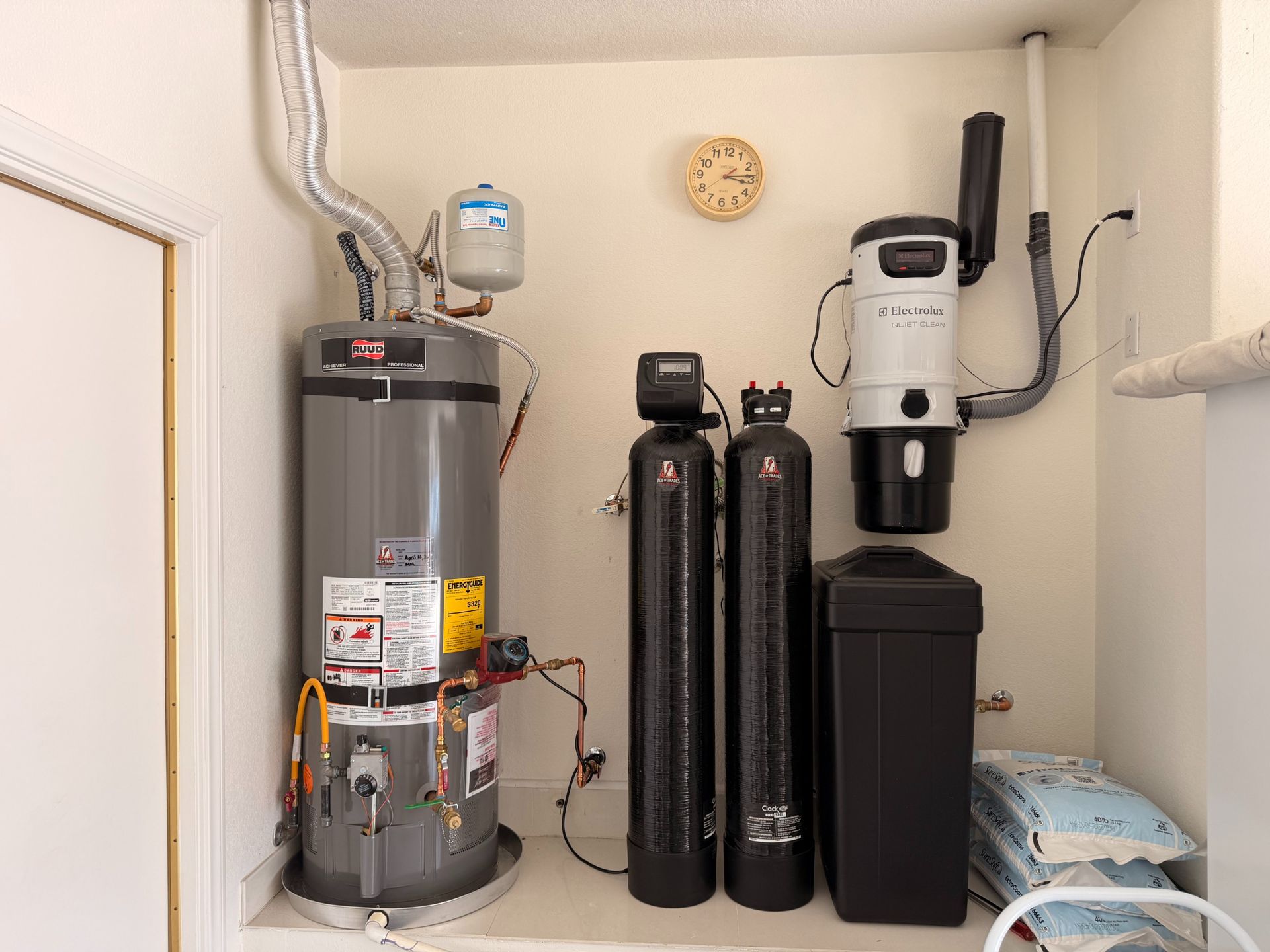 Water heater and water softener system in a small utility closet with pipes, tanks, and a filter unit.
