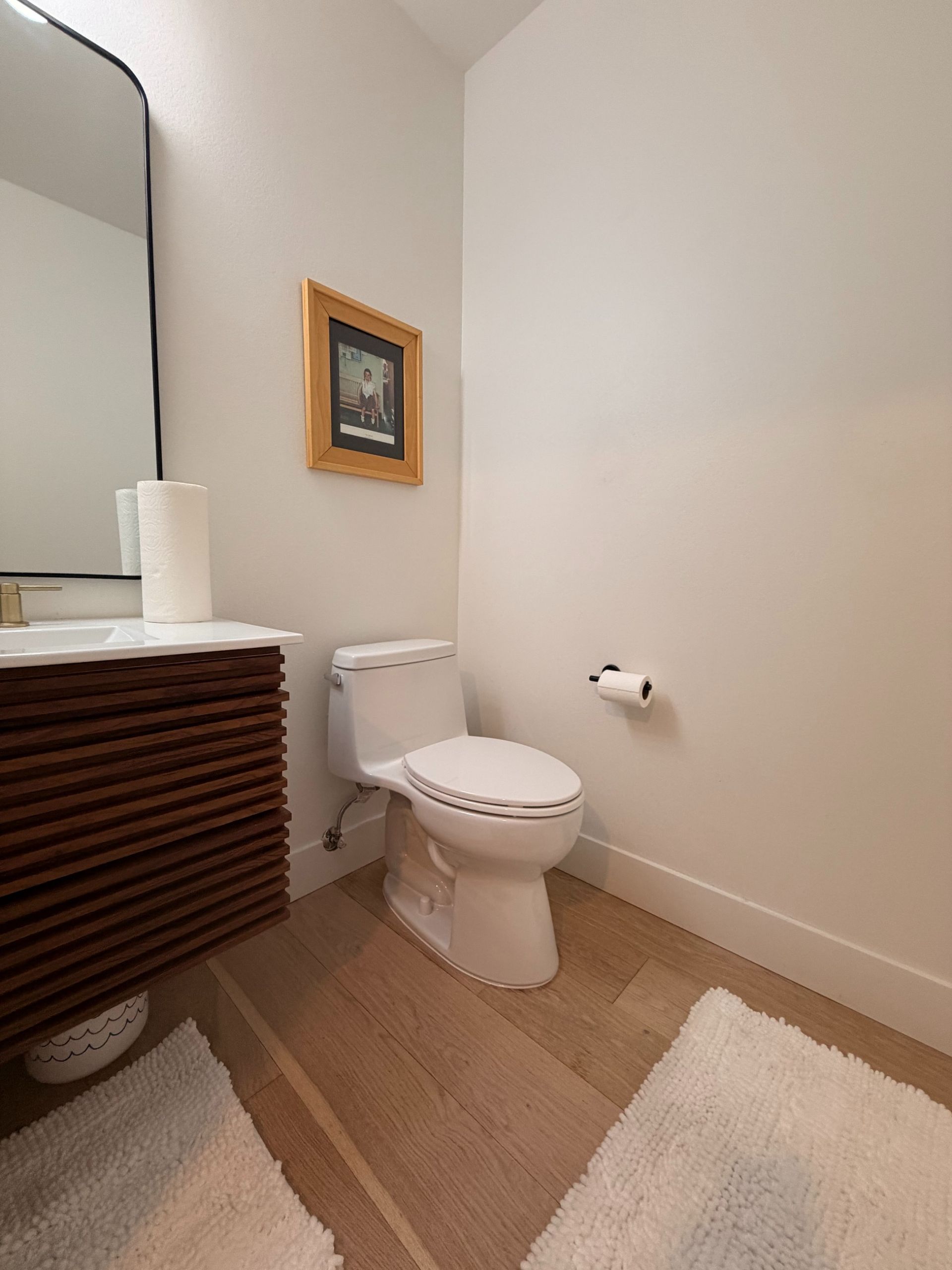 Small bathroom with toilet, sink cabinet, wall art, and beige rugs.