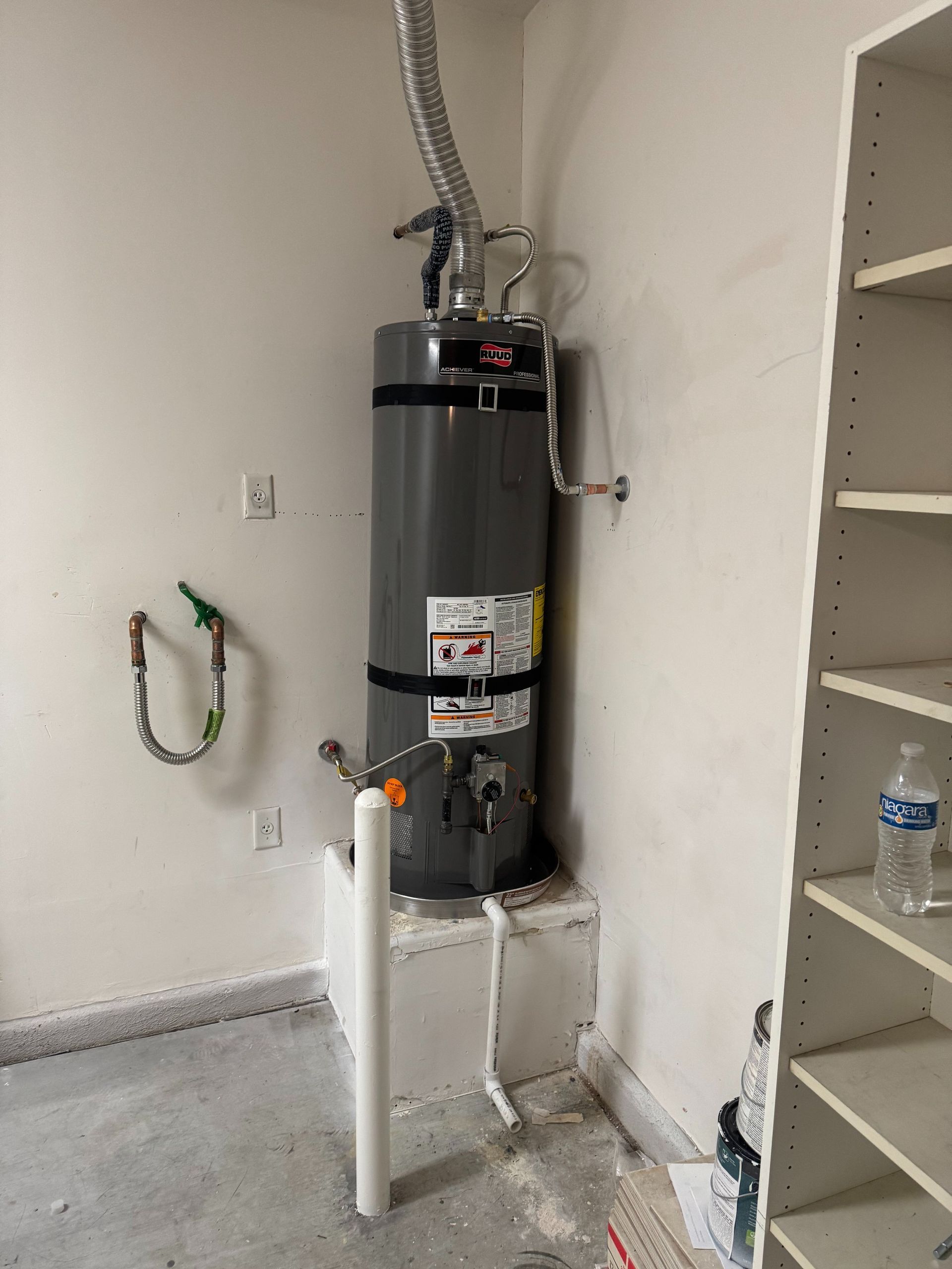 Water heater in a small utility room with exposed pipes and nearby shelving.