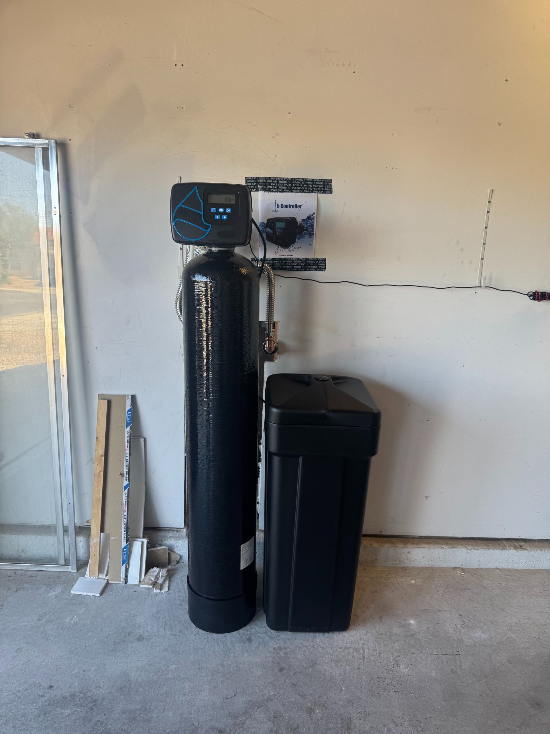 Water softener and filtration tanks beside a wall in a basement or utility room.