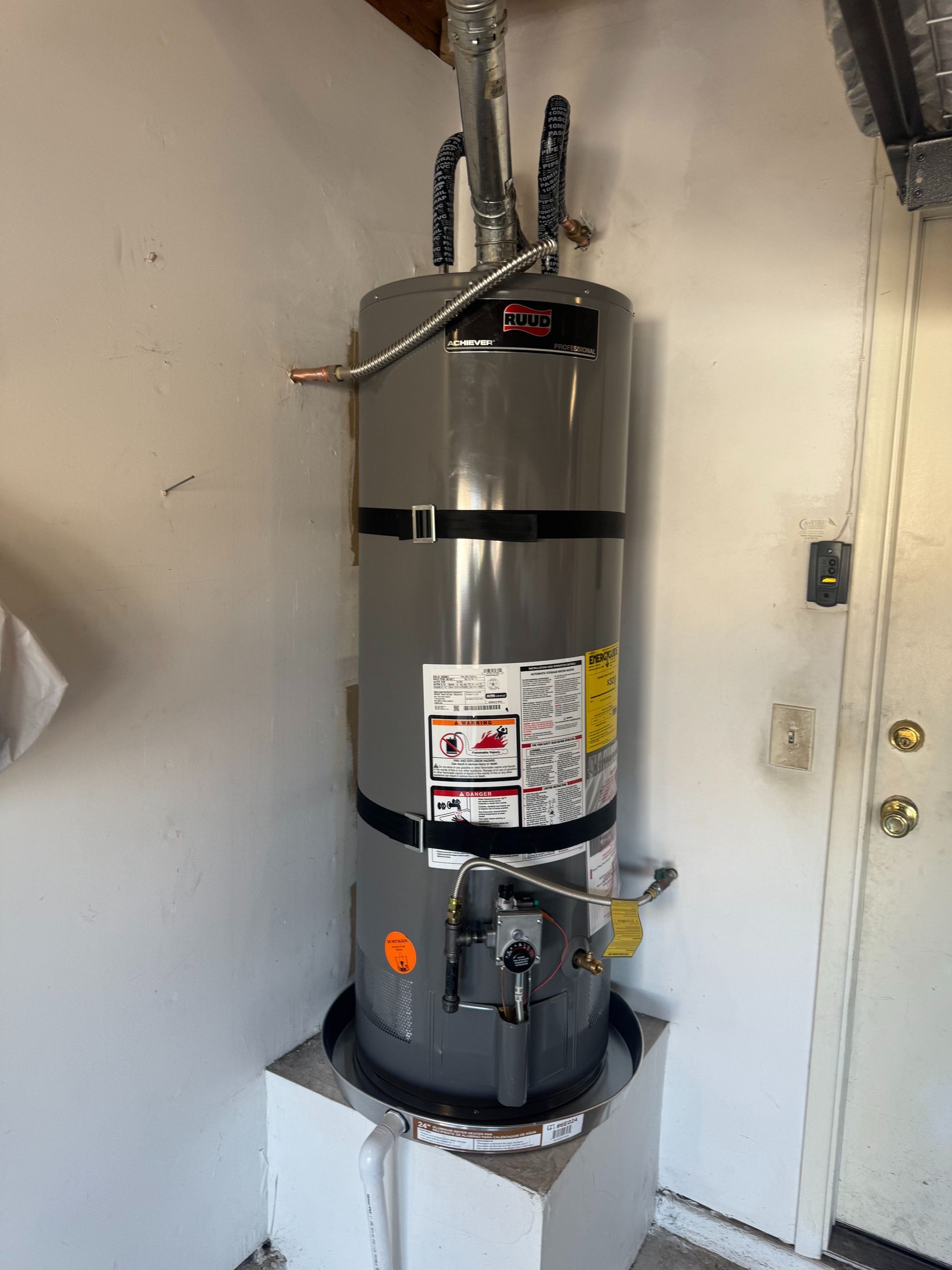 Tall gray water heater in a narrow utility closet with pipes and labels on the front.