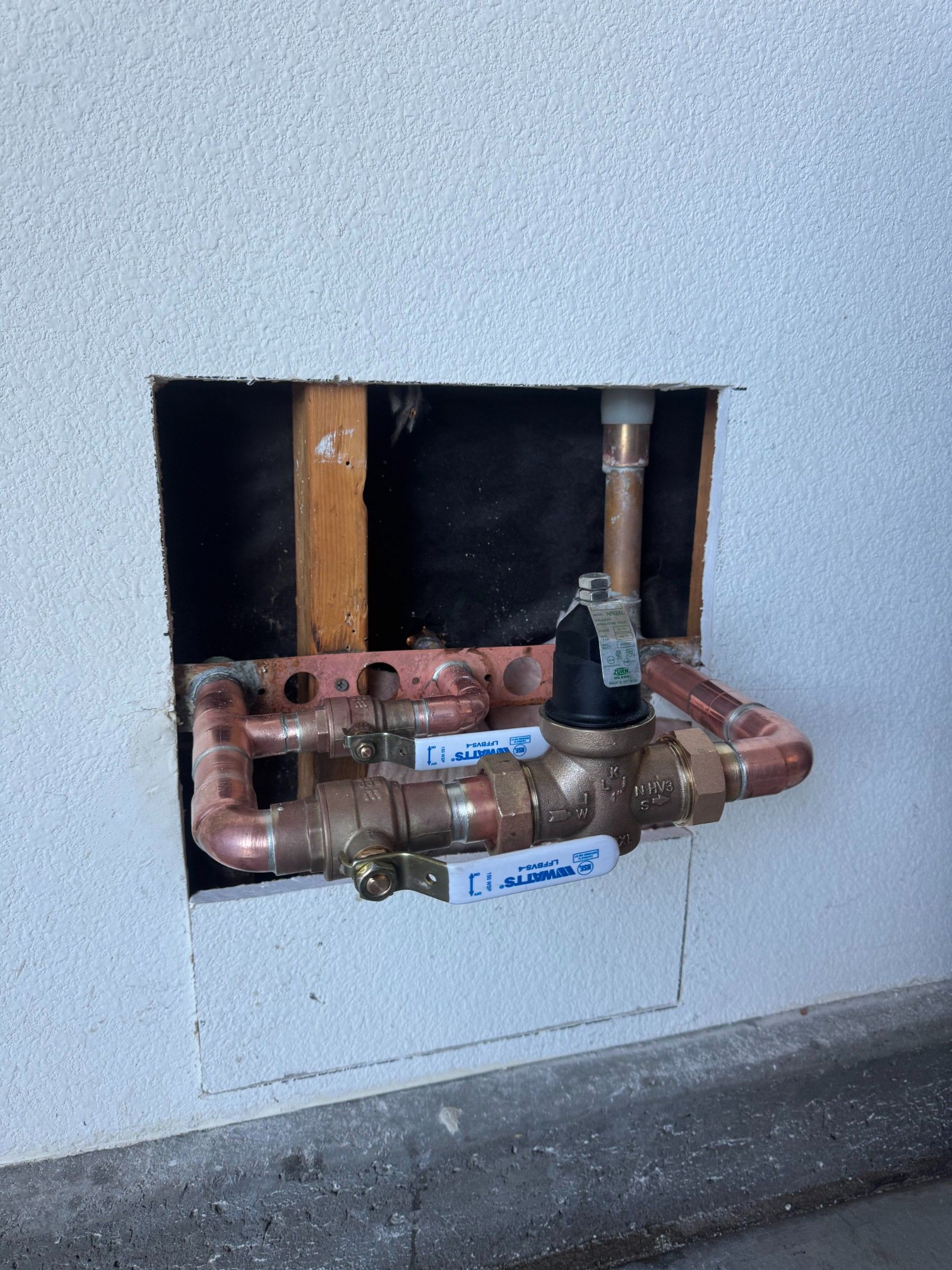 Exposed plumbing and shutoff valves inside a wall opening, with copper pipes and a wooden stud visible.
