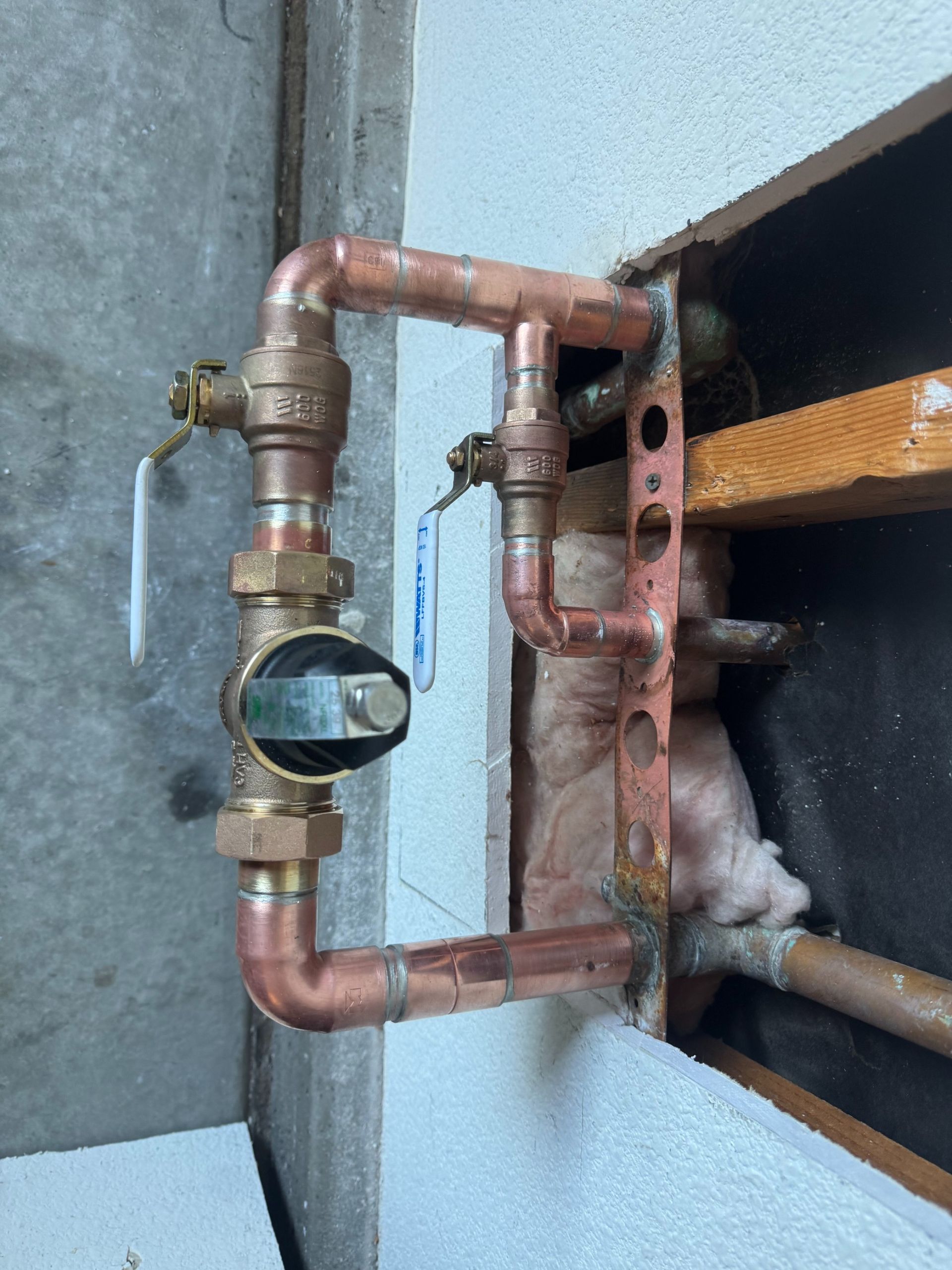 Copper pipes and brass water meter installed in a wall opening with exposed insulation and framing.