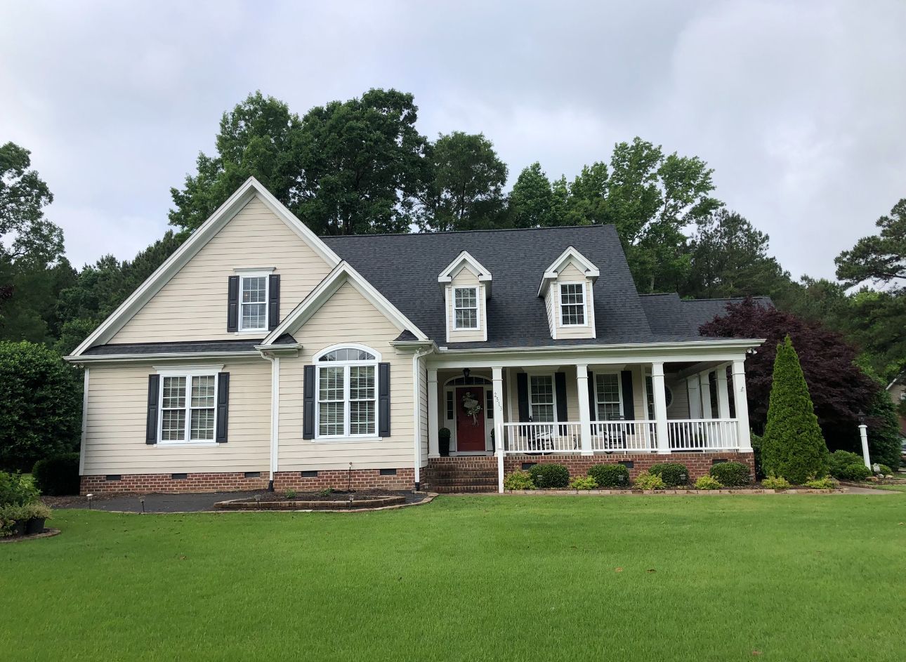 Cream-colored suburban house with a front porch, green lawn, and trees under a cloudy sky