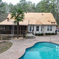 House with tan roof, front porch, and turquoise swimming pool in a wooded yard