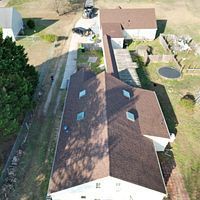 Aerial view of a house with a brown roof, driveway, and landscaped backyard with a circular pond.
