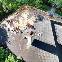 Workers repairing a shingled roof around two chimneys, seen from above.