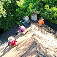Workers on a shingled roof repairing it, surrounded by green trees.
