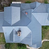 Aerial view of a gray metal roof with multiple peaks, vents, and a chimney, bordered by grass.