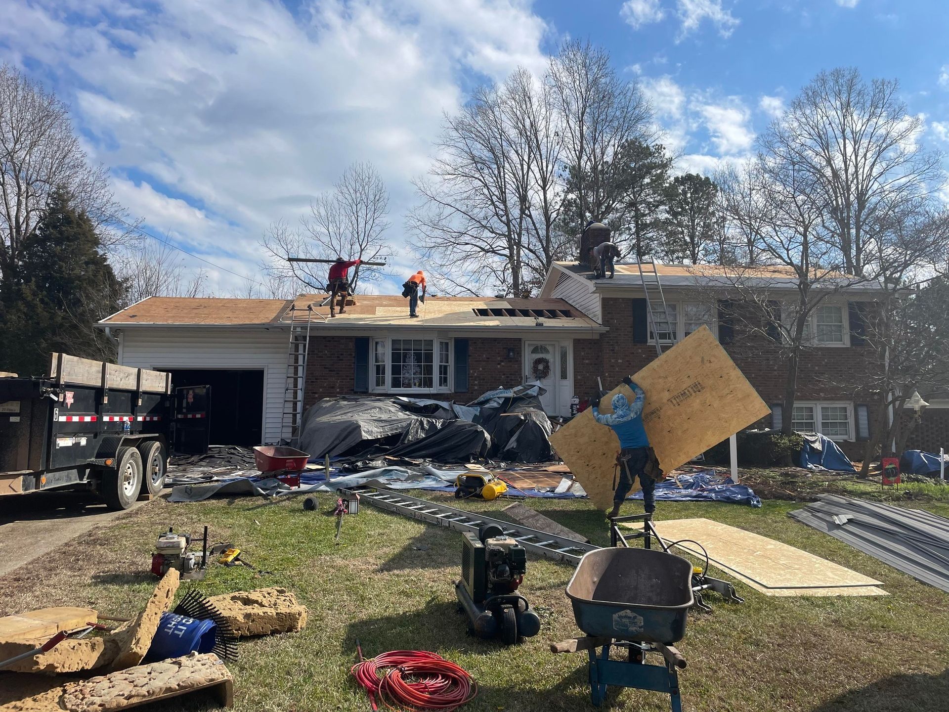 Storm-damaged house with workers clearing debris and a roof repair in progress in the yard