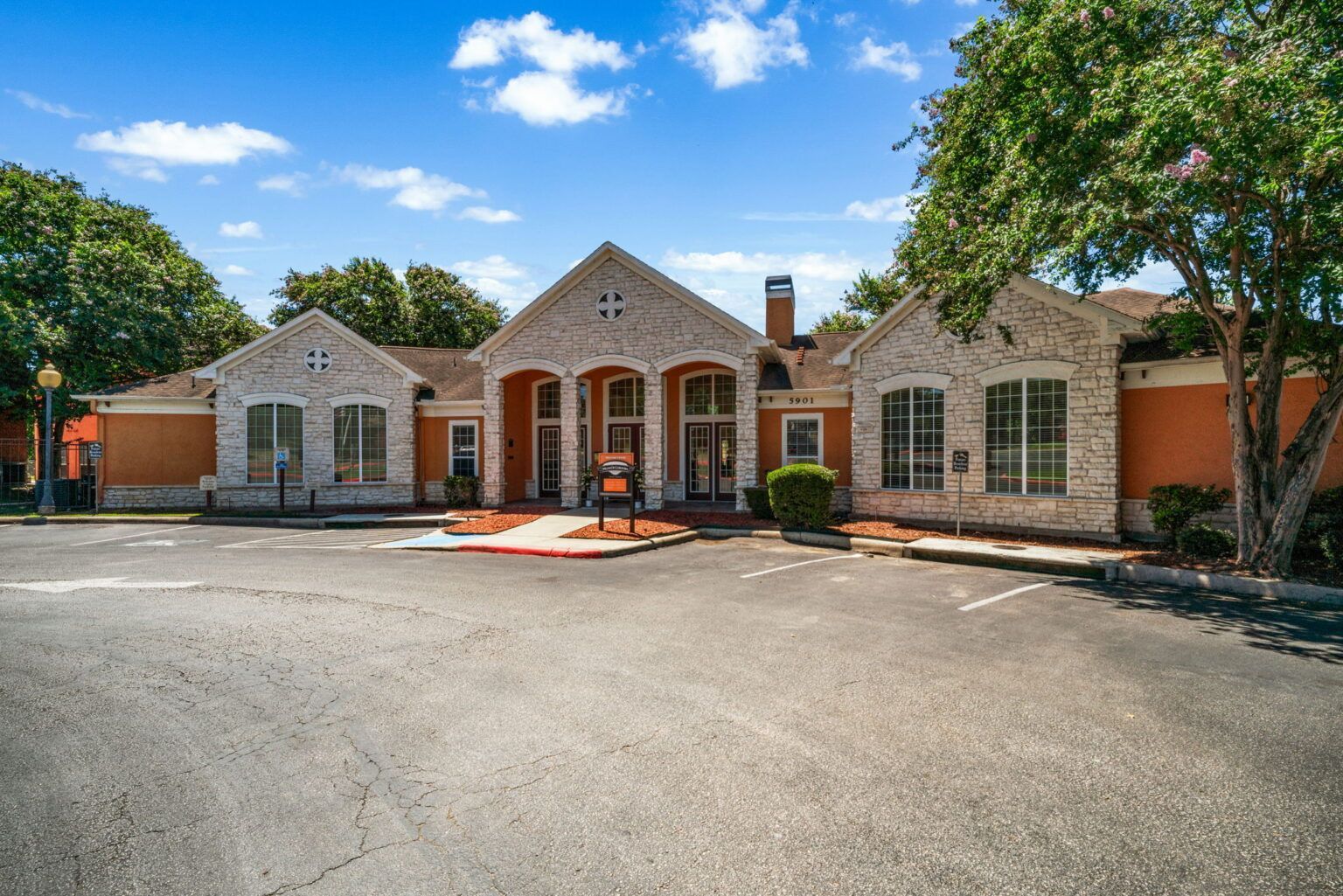Front view of a stone-and-brick building with arched entry, red doors, and a wide paved driveway under a blue sky