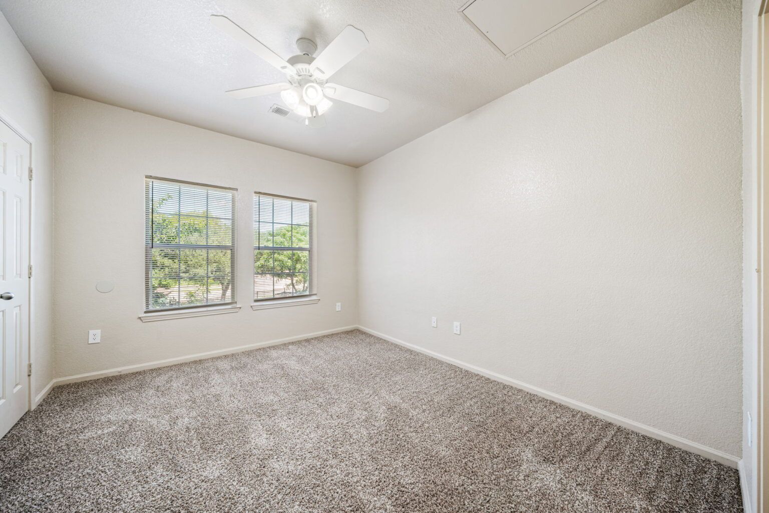 Empty beige bedroom with carpet, white ceiling fan, and two windows letting in natural light