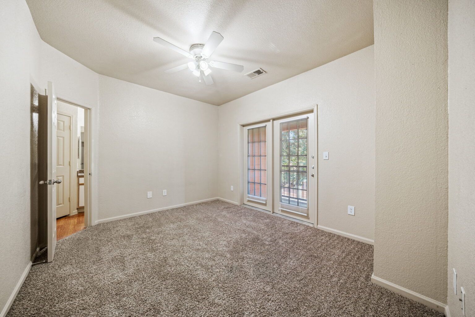 Empty carpeted bedroom with white walls, ceiling fan, and glass doors to a balcony.