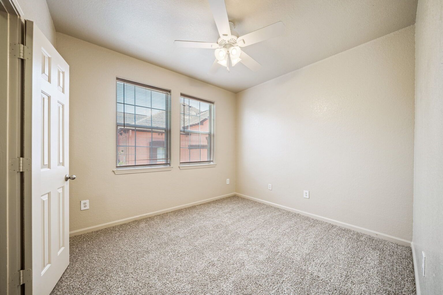 Empty beige bedroom with carpet, two windows, and a ceiling fan