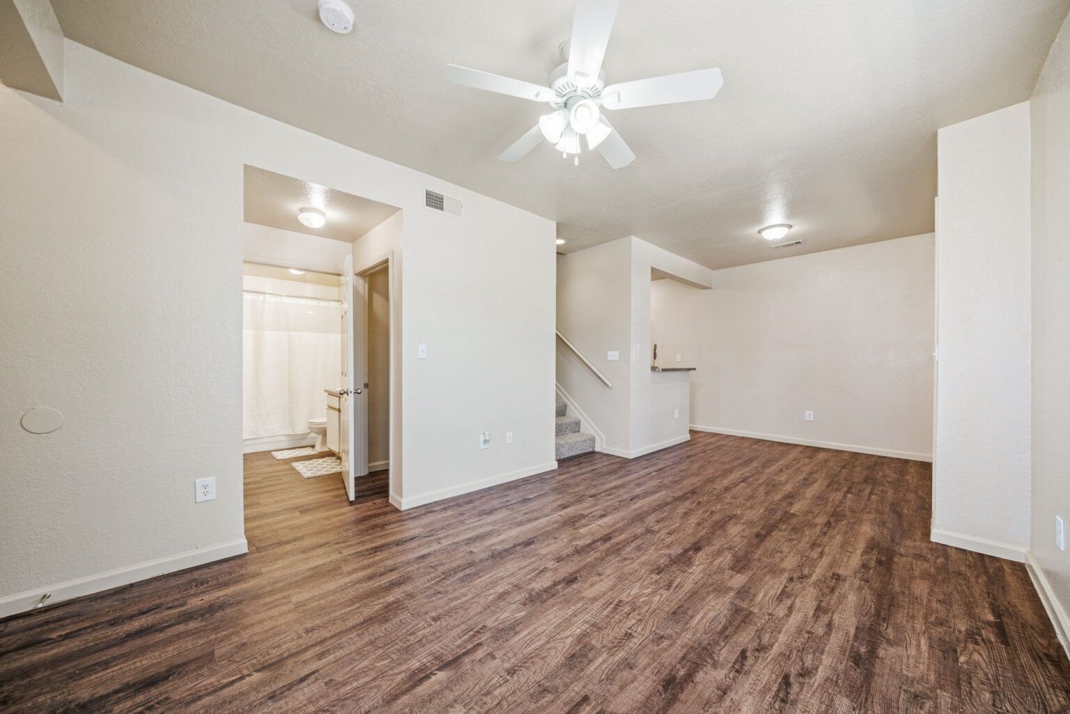 Empty beige living room with wood-look flooring, ceiling fan, and open doorway to kitchen.