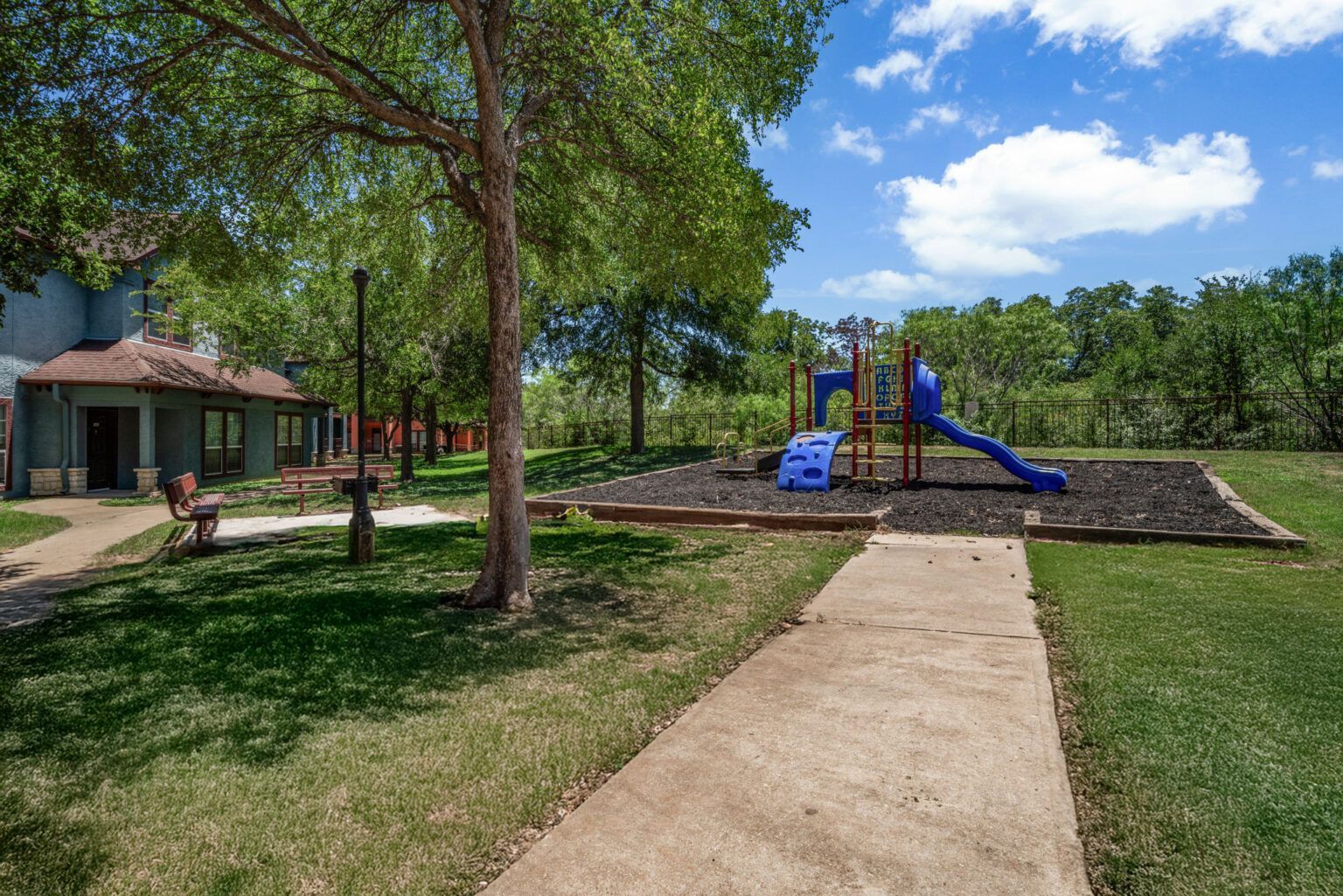 Park path leading to a playground with blue slide, trees, and a pavilion on a sunny day