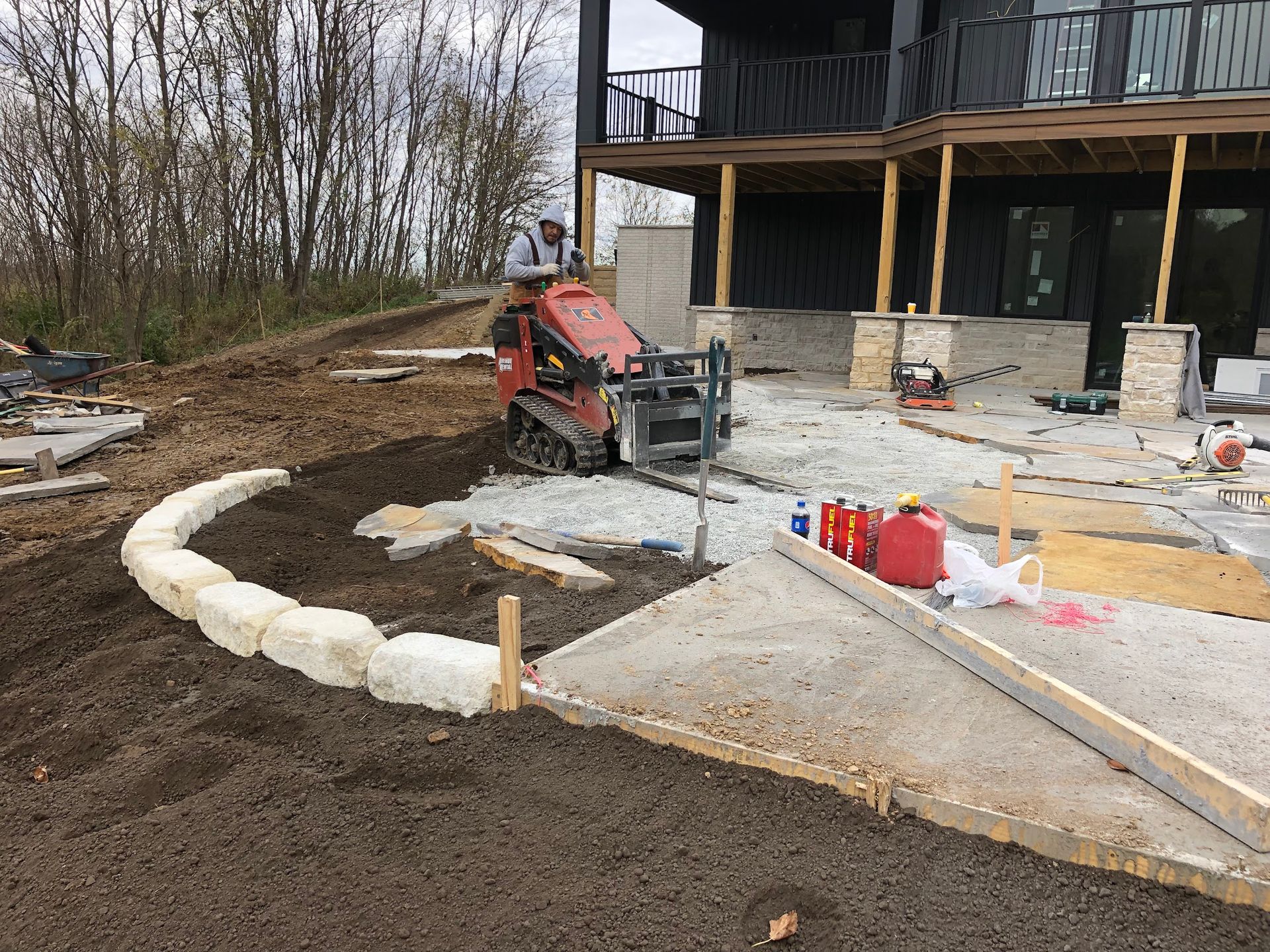 Man operating mini skid steer on construction site with patio and house.