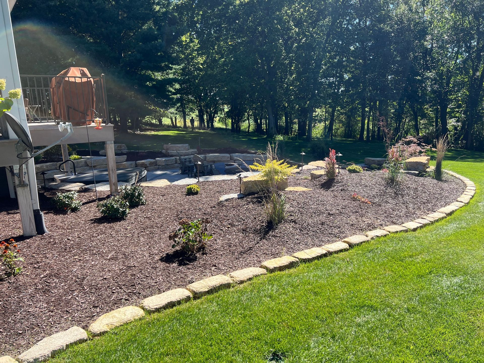 Landscaped yard with dark mulch, plants, and stone edging, adjacent to a deck and green lawn.