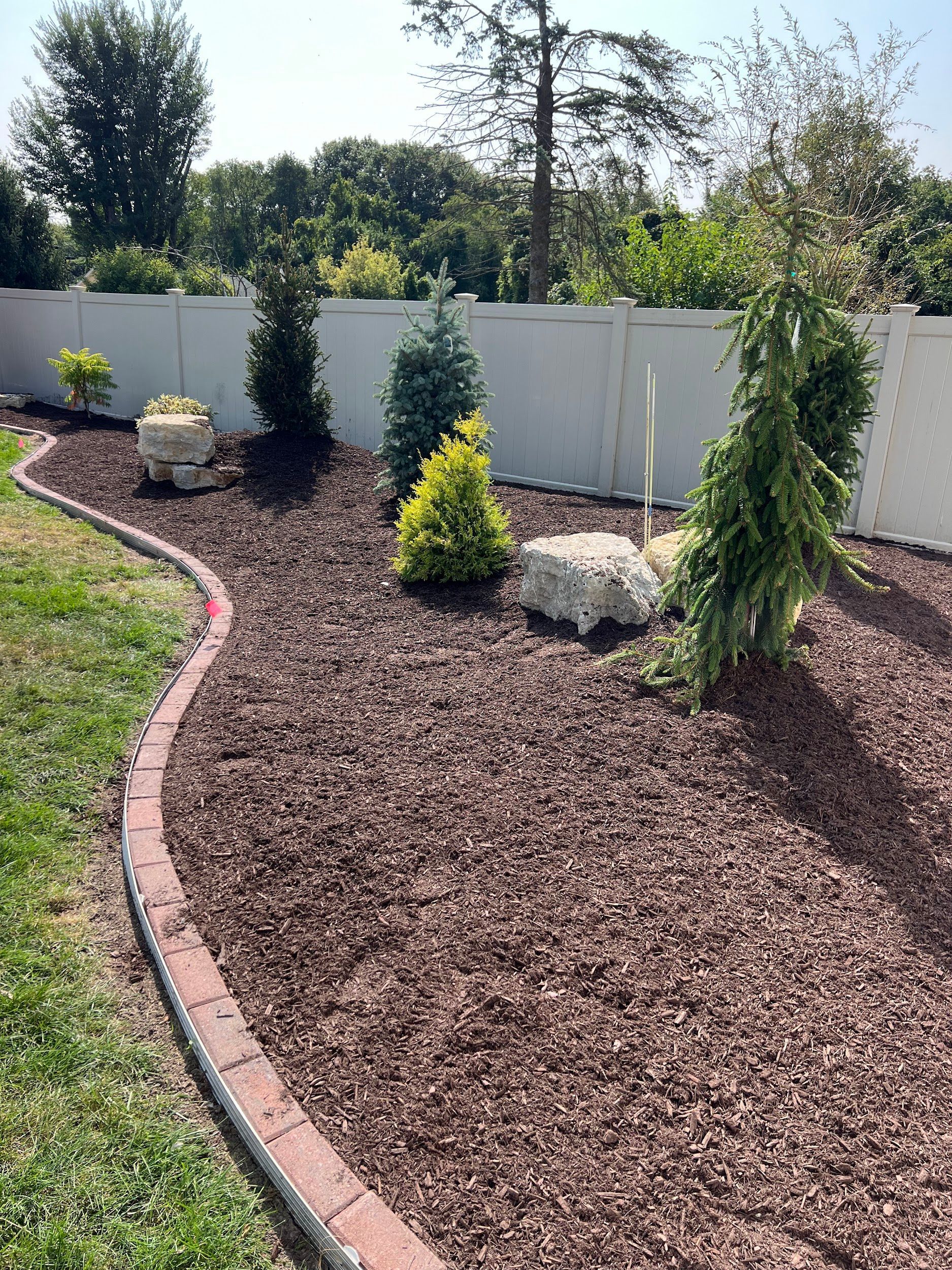 A landscaped garden bed with brown mulch, small trees, and a decorative brick border alongside a white fence.