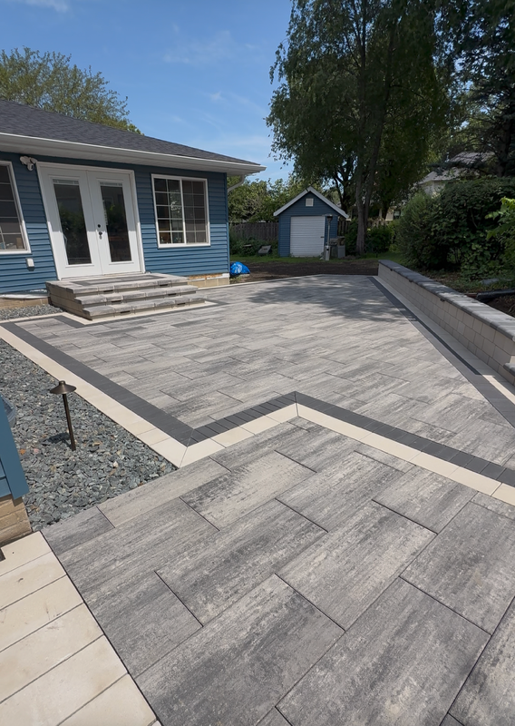 Brick patio with decorative pattern, brown border, in front of a house with steps and door.
